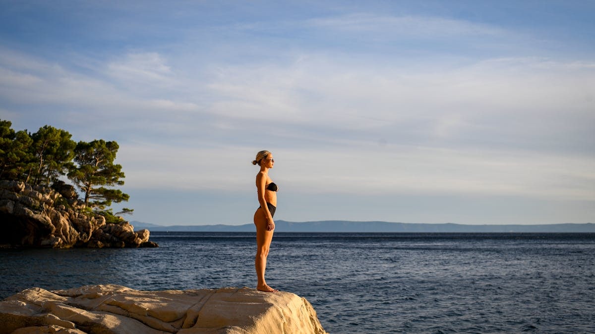 A woman in a black bikini enjoys a serene sunset on a rocky cliff by the Adriatic Sea in Croatia.